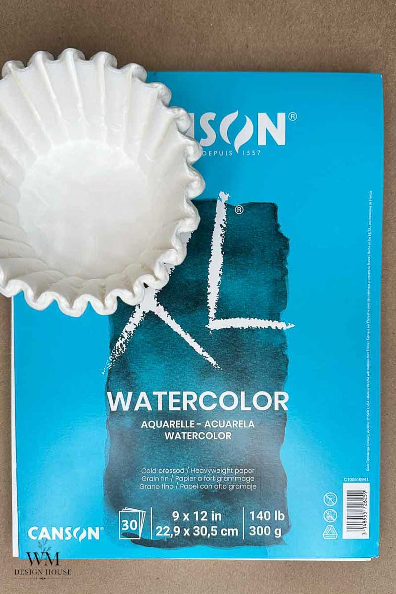 A pack of Canson watercolor paper lies on a brown surface beside a white coffee filter, evoking curiosity about how to make coffee stained paper. 