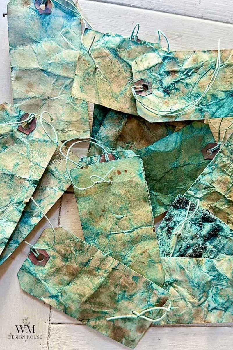 A pile of handmade paper tags with marbled green, blue, and brown patterns, some featuring a subtle coffee stained paper effect, each with a string and a hole reinforced by a metal ring. The tags are spread out on a light wooden surface.