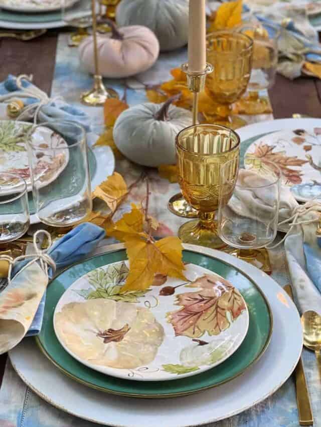 Place setting at a fall harvest table. A cream-colored charger plate with a green dinner plate and a pumkin design on the salad plate. Amber glasses and gold silverware accent the table nicely.