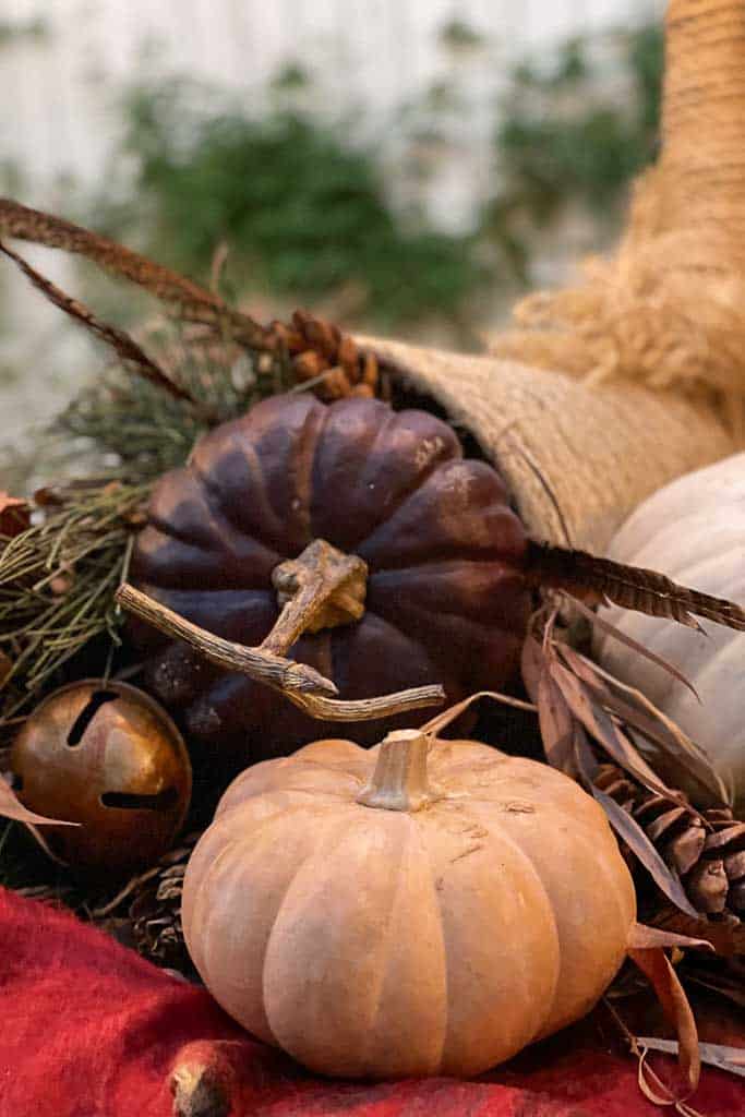 An autumn-themed arrangement features a small beige pumpkin, a dark purple gourd, and a white pumpkin, surrounded by dried leaves, twigs, and a brass bell, all spilling out of a woven DIY cornucopia rope basket. The background is softly blurred greenery.