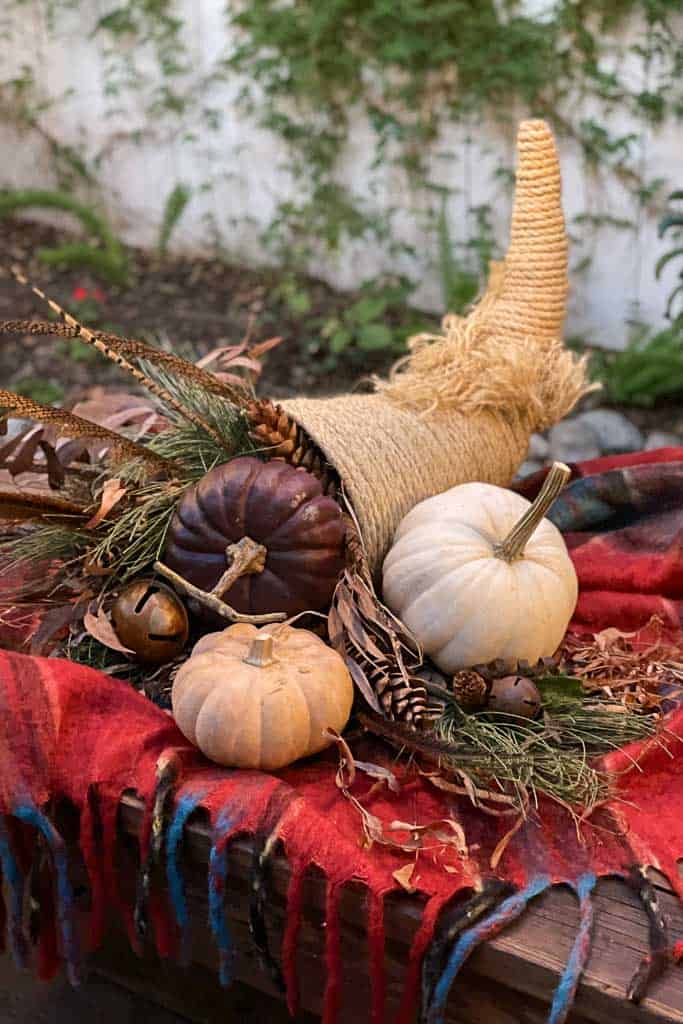 A rustic DIY rope cornucopia is displayed outdoors, filled with pumpkins of various colors, pinecones, acorns, and dried foliage. The cornucopia rests on a red and multicolored plaid blanket, with a natural background of greenery and a white fence.