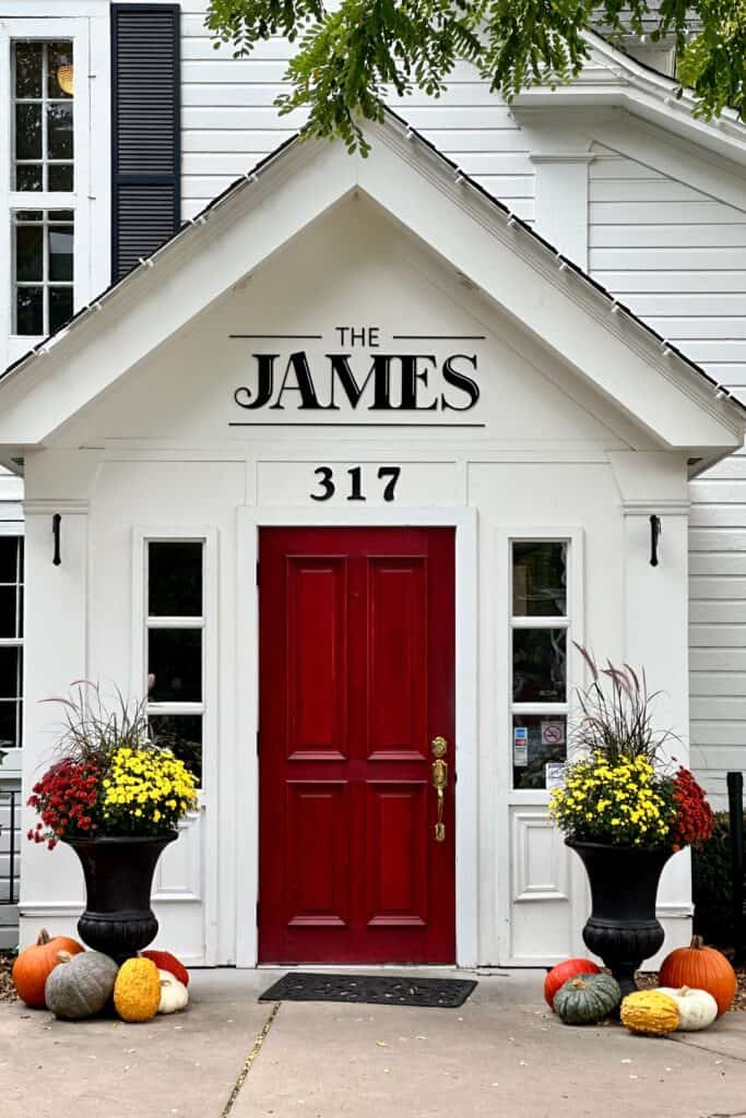White charming house with a red door and two beautiful planters on either side of the door. It is now a restaurant with the name James above the door. 