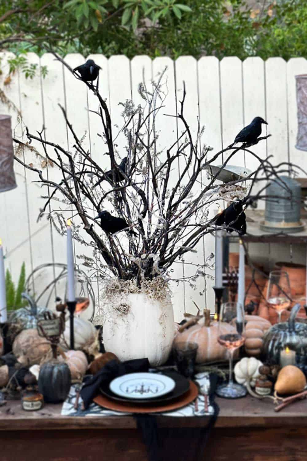 A white pumpkin vase acts as a DIY Halloween tree, holding bare branches with black artificial crows perched on them, surrounded by candles, pumpkins, and tableware for a spooky Halloween-themed tablescape.