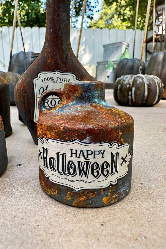 Rustic Halloween-themed jug with a "Happy Halloween" label, featuring a weathered appearance. In the background are similarly aged bottles and a pumpkin, set on a light floor with a white fence and greenery behind.