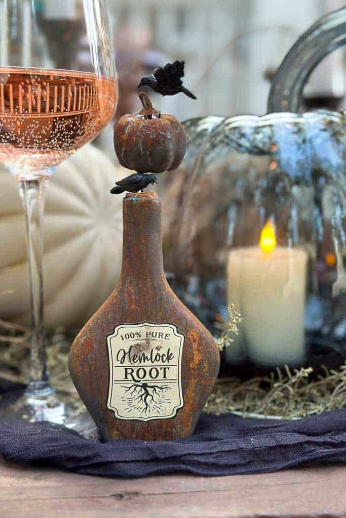 A rustic, antique-style bottle labeled "100% Pure Hemlock Root" is displayed on a dark cloth. The bottle's stopper is adorned with a small black bird and a miniature pumpkin. In the background, a glass of rosé wine and a lit candle inside a glass pumpkin are visible.