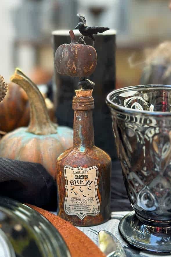 A Rusty mini liquor bottle that has been rusted with a Bats Brew label on the front. A small pumpkin and a black crow are placed on the top for decoration. 