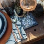 A rustic table setting with a wooden surface features a black-rimmed plate with an intricate design, a dark glass goblet, a spoon and fork with wooden handles, and a glass of rosé wine on an ornamental resin coaster. Decorative elements include moss, pumpkins, and dried flowers.