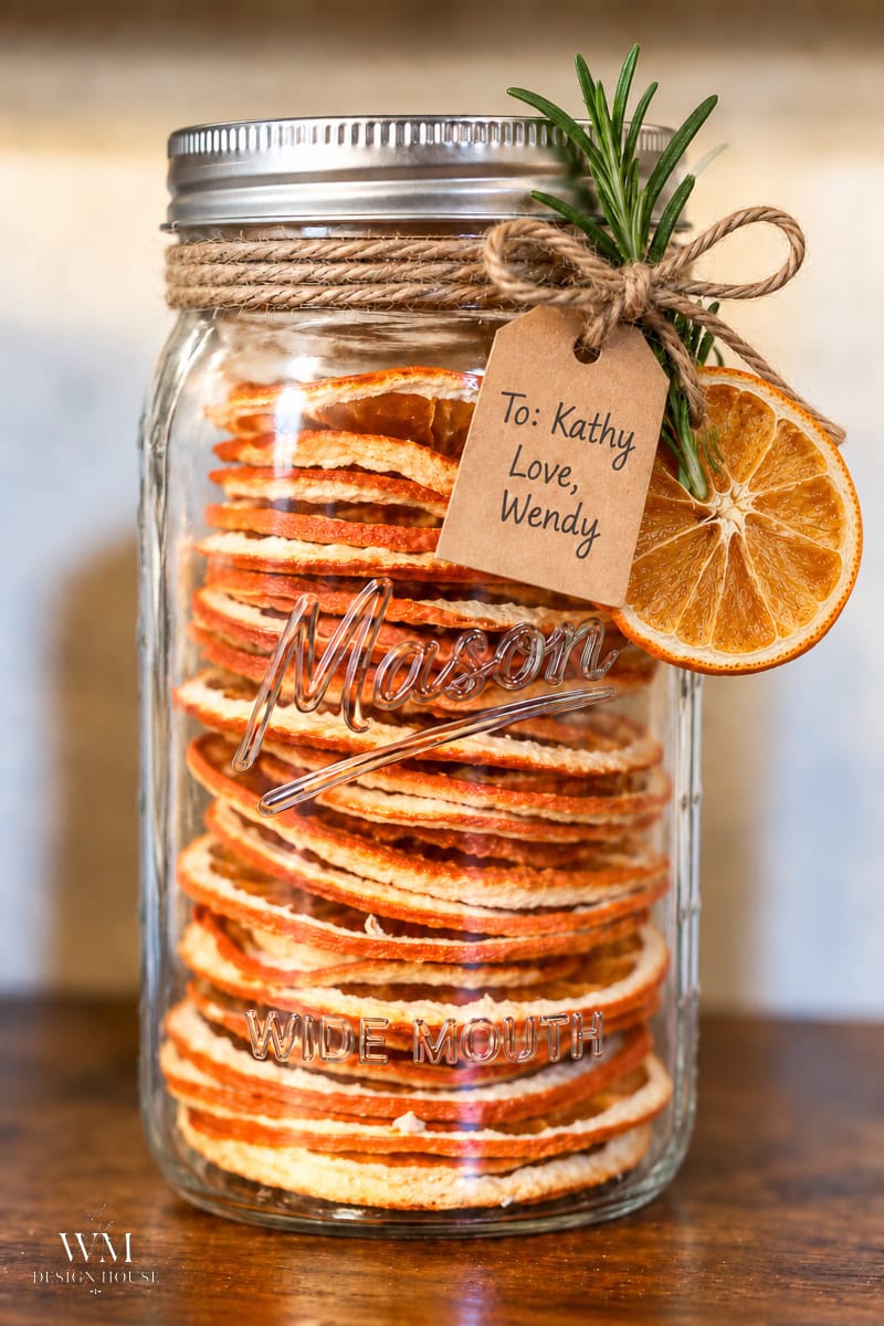 A Mason jar filled with dried orange slices, tied with twine and a sprig of rosemary, featuring a gift tag that reads “To: Kathy, Love, Wendy.”.