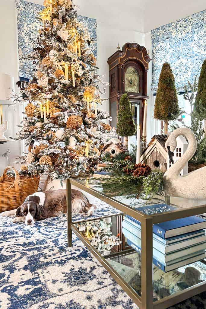 A view of a blue and white living room with a natural Christmas theme. The coffee table is decorated with moss trees, wood birdhouses, greenery, and a swan. The natural white flocked Christmas tree is decorated with dried hydrangeas, white ornaments, and hanging candles.