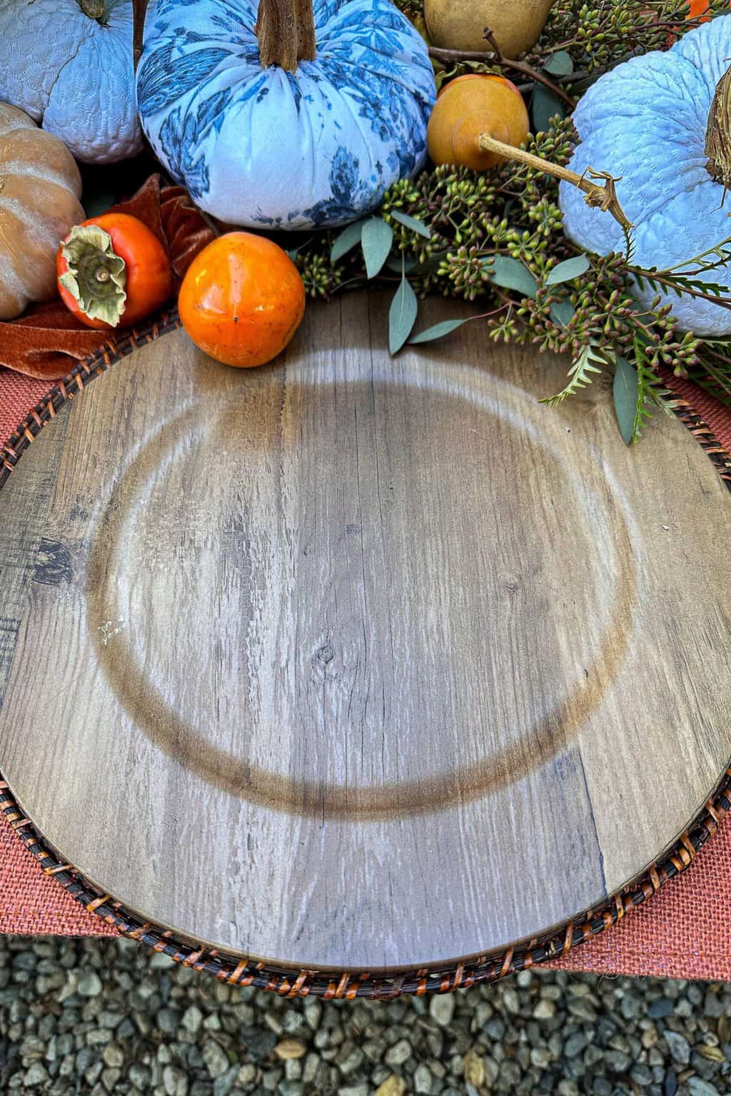 Table setting in progress with two chargers sitting on a table decorated with persimmons and blue and white pumpkins.
