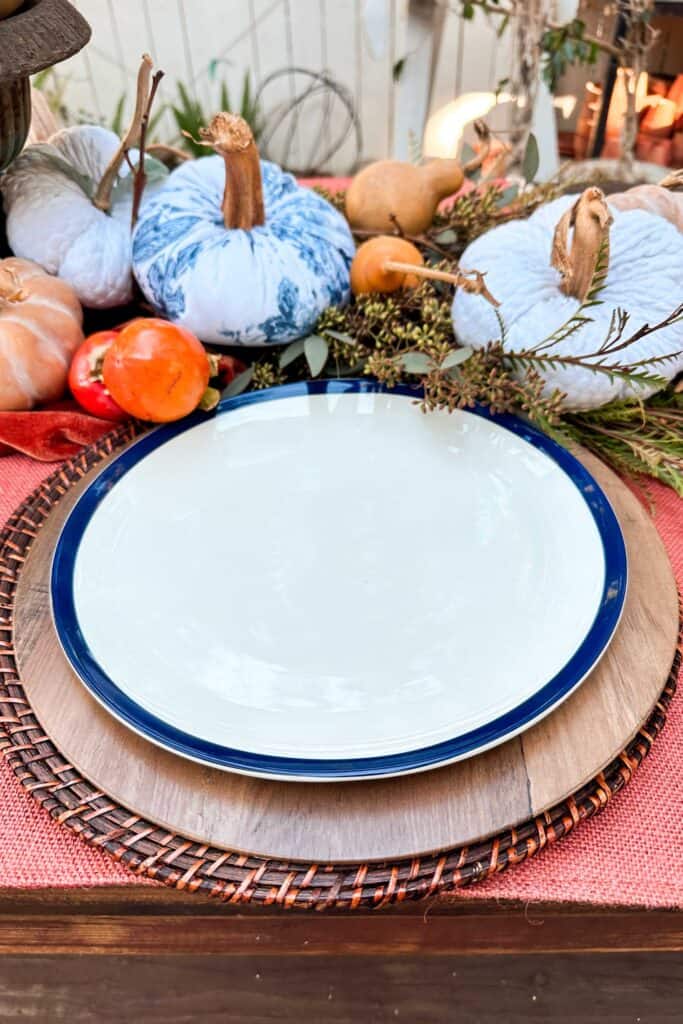 Table setting in progress with two chargers and a dinner plate sitting on a table decorated with persimmons and blue and white pumpkins.