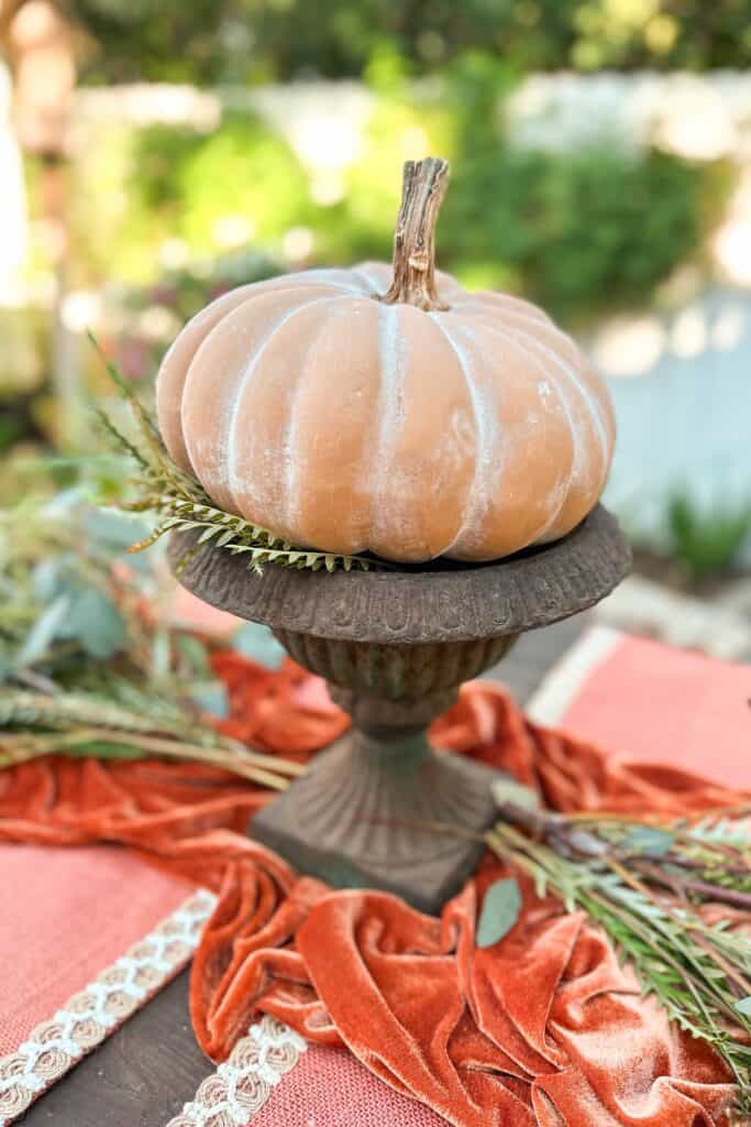 Pumpkin centerpiece sitting on top of a vintage urn on a table with a rust-colored table runner.