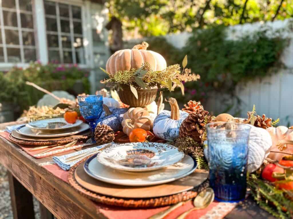 A wooden table is set outdoors with festive fall decor including a centerpiece of a pumpkin on a pedestal, assorted pumpkins, and pinecones. The blue and orange Thanksgiving table ideas come to life with plates, blue glasses, and napkins surrounded by greenery and autumn leaves.