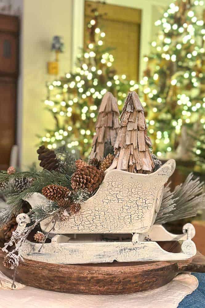 A miniature wooden vintage sleigh painted white filled with greenery, pinecones, and wooden trees.
