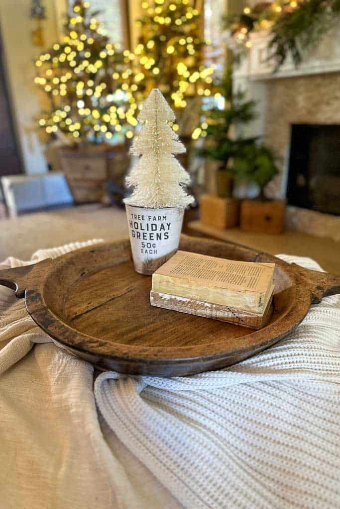 A wood tray sitting on an ottoman in the family room in the process of creating a vignette for Christmas. I have started with a few old books stacked for height and a tree in a holiday greens container.