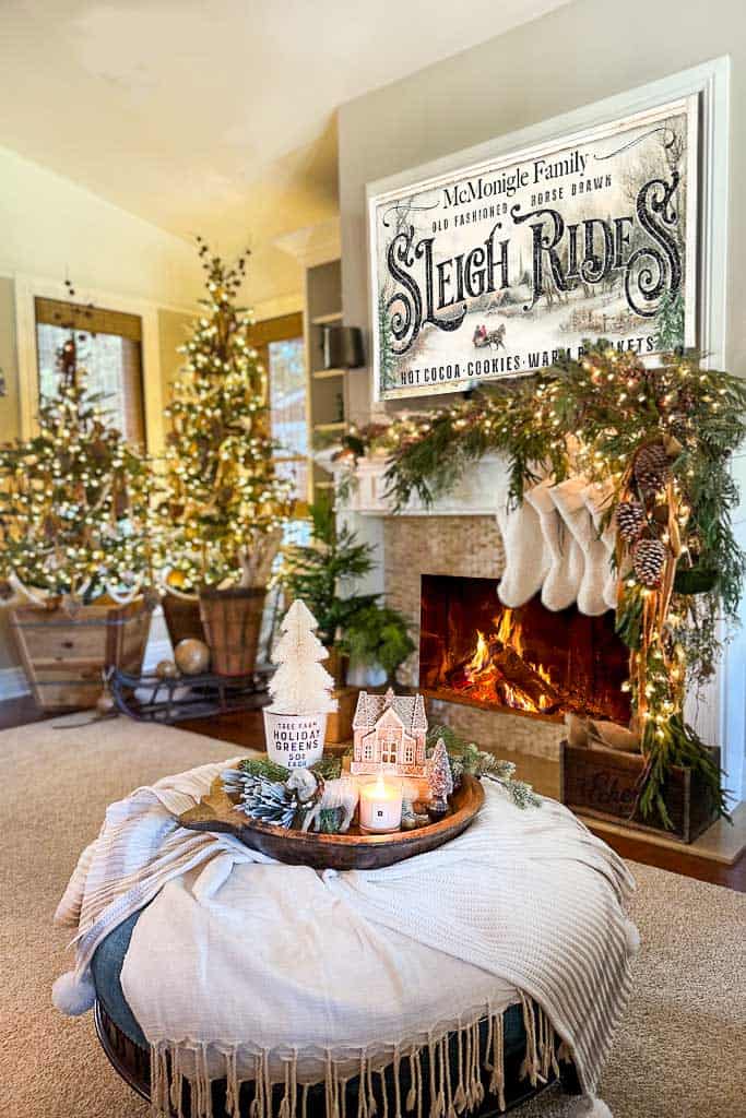 A family room with Two Christmas trees, a fireplace with stockings, and a sign hanging above the fire that says sleigh rides. The coffee table is draped with blankets and a tray rilled with Christmas decor.