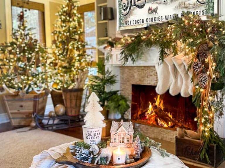 A family room with Two Christmas trees, a fireplace with stockings, and a sign hanging above the fire that says sleigh rides. The coffee table is draped with blankets and a tray filled with Christmas decor.