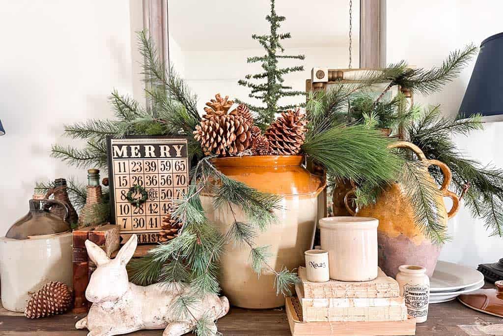 A side bar in the dining room is decorated with natural colors and elements for the holidays. Lots of fresh greenery, pinecones, and natural pottery layered with old books and crocks.