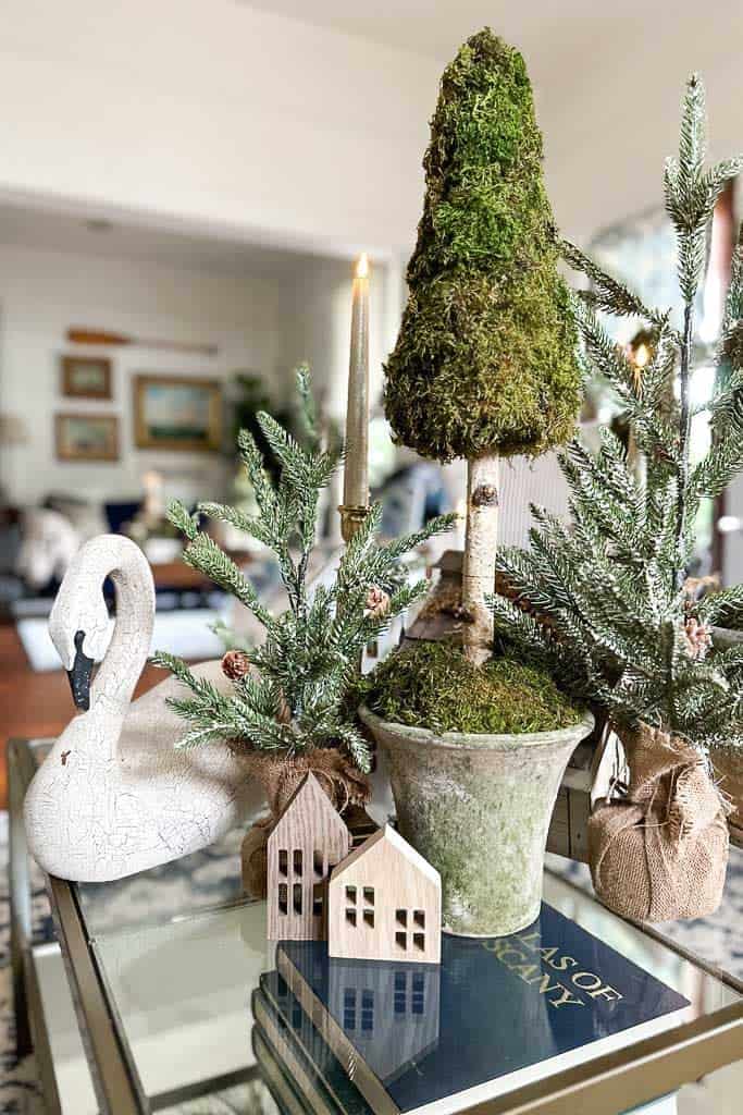 An up-close image of a glass coffee table that is decorated for Christmas with a moss Christmas tree, fresh greenery, a few wood houses, and a white swan.