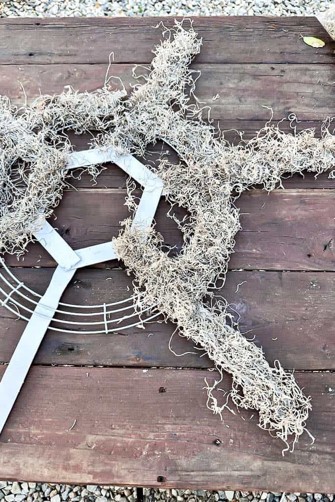 Wire wreath frame with wooden snowflake sticks that are being mossed before adding pinecones.