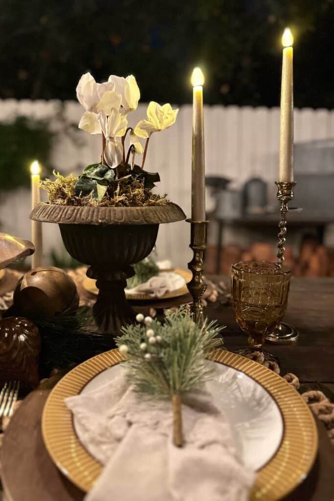 Place setting for a rustic Christmas table including a rope placemat, wood chargers, god and white plates with a linen napkin, and a sprig of greenery with gold flatware.