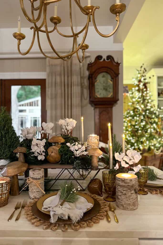 The indoor dining room table is set with a rustic tablescape, including a wooden sled for a centerpiece, with wood mushrooms and ornaments sitting on top of the sled amongst white cyclamen and a rosemary Christmas tree.