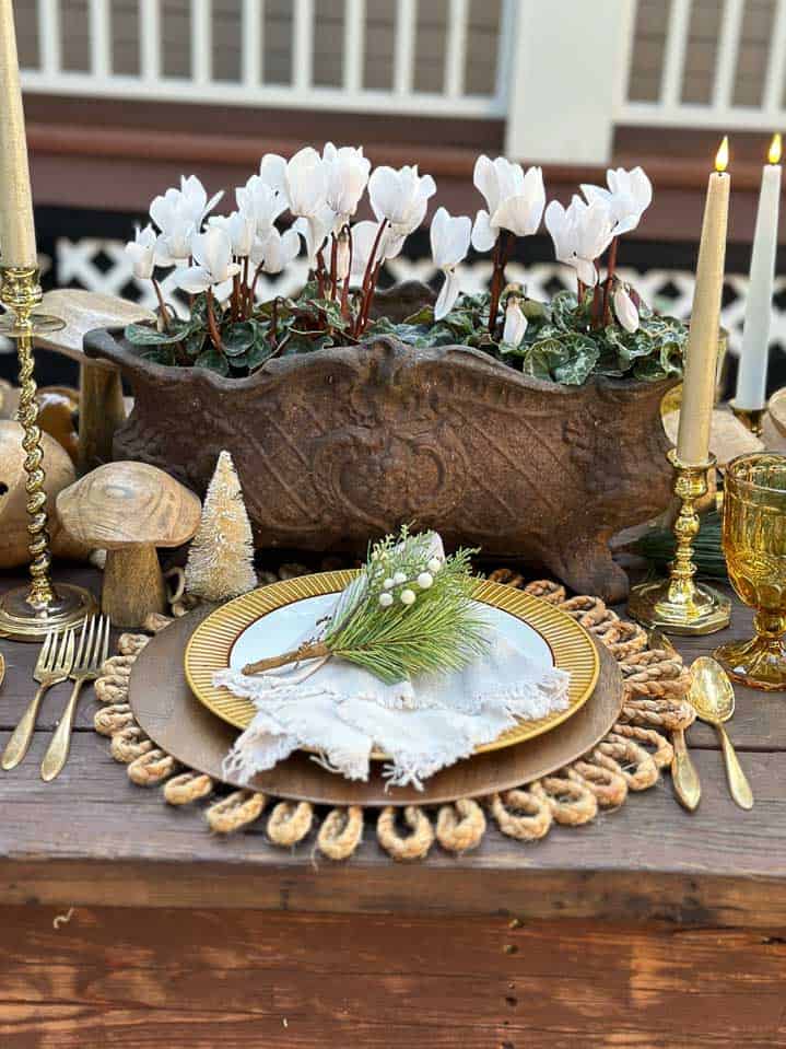 Place setting with a wood charger and gold and white plates with a natural linen napkin and fresh greenery on top.