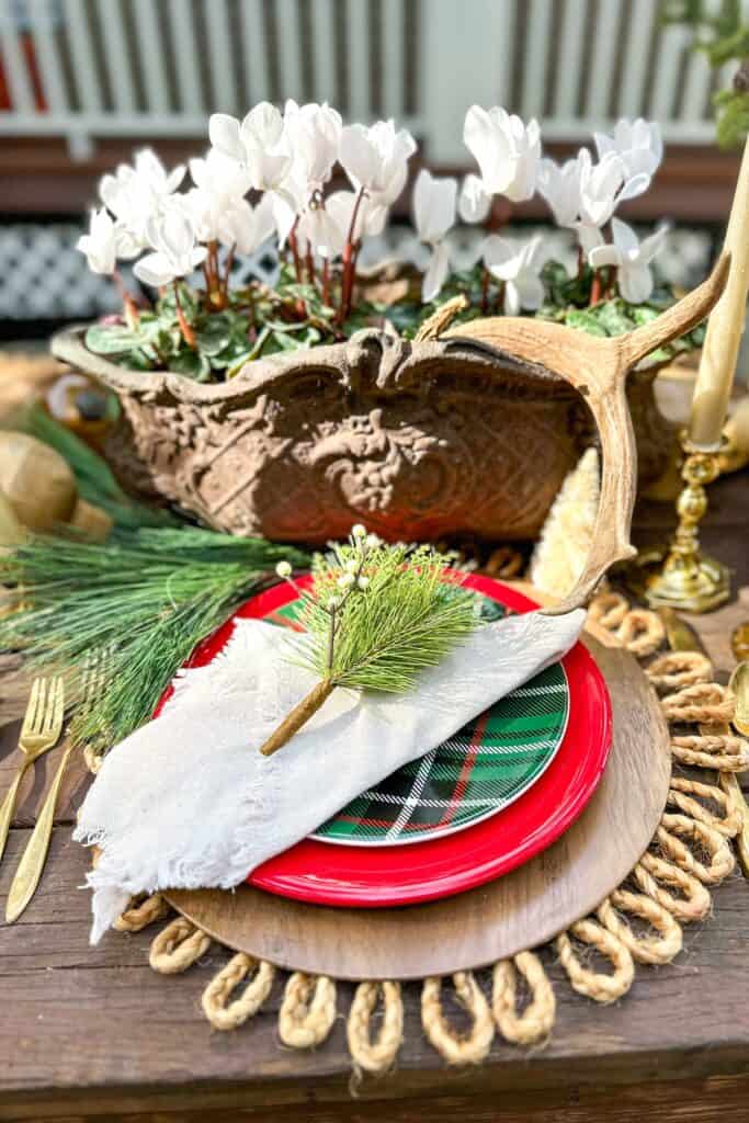 Red and plaid plates layered on a wood charger plate and a rope placemat to create a rustic tablescape for Christmas. A vintage urn filled with white cyclamen and antlers is the centerpiece of the table.