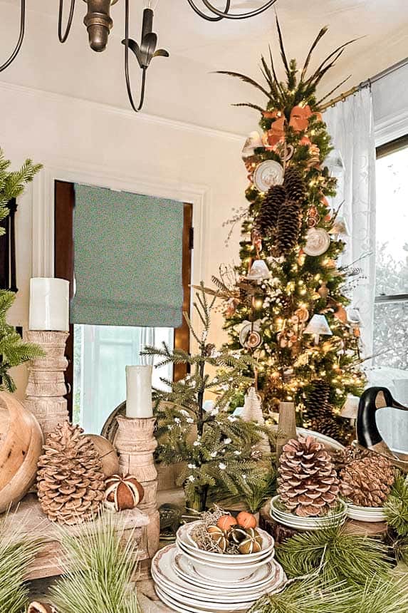 The dining room table is decorated with a wooden goose, greenery, and wood candlesticks. There is a stack of vintage plates on the table with dried oranges and pinecones. A neutral Christmas tree sits in the back. 