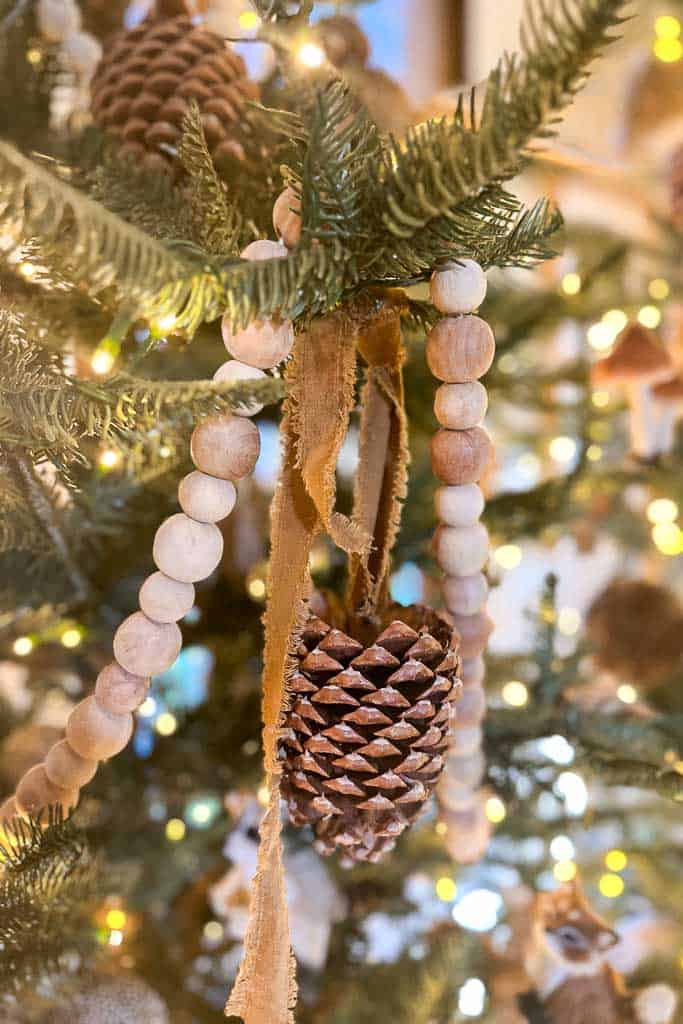 A close-up of a Christmas tree decorated with string lights, wooden bead garlands, and a pinecone ornament tied with a rustic ribbon. The pine branches are lush and green, creating a festive and cozy atmosphere.