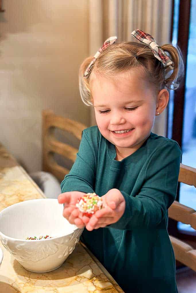 A little girl rolling cookie dough into a ball and then covering it with sprinkles.