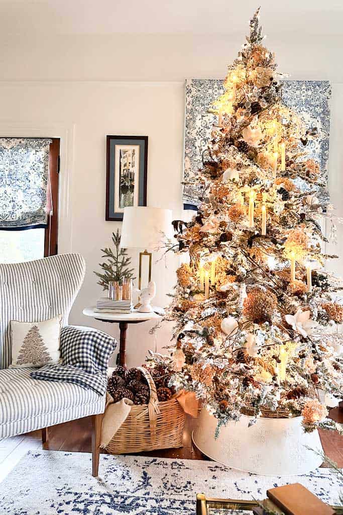 A cozy living room features a large, neutral Christmas tree adorned with lights, pinecones, and ornaments. A blue-striped armchair with a small tree pillow sits next to a wicker basket filled with pinecones. A side table holds a small tree and lamp.