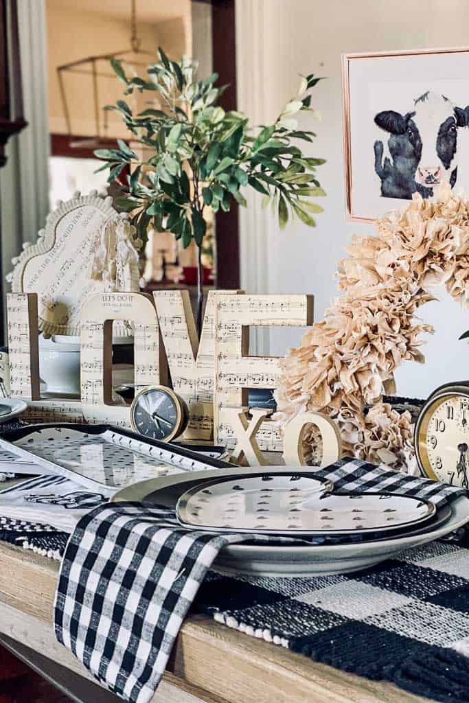 A rustic-themed dining table is decorated with a black-and-white checkered table runner, plates, a plant, vintage clocks, and a wooden "LOVE" sign covered in sheet music. Perfect for Valentine's Day Dinner Table Decoration Ideas, cow-themed artwork and an "XO" decoration add extra charm to the setting.