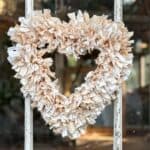 A DIY Rag Wreath, heart-shaped and crafted from beige and white fabric strips that were dyed with tea. The wreath hangs on a rustic, weathered windowpane with a blurred indoor background.