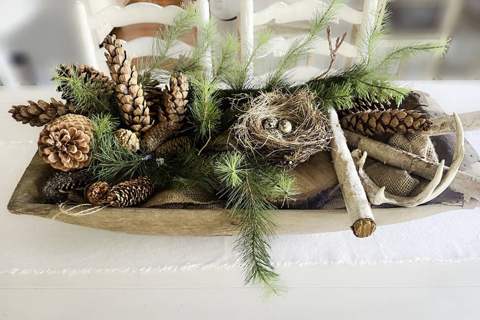 A wood dough bowl filled with greens, pinecones, birch logs and more. Winter decorations (not Christmas).