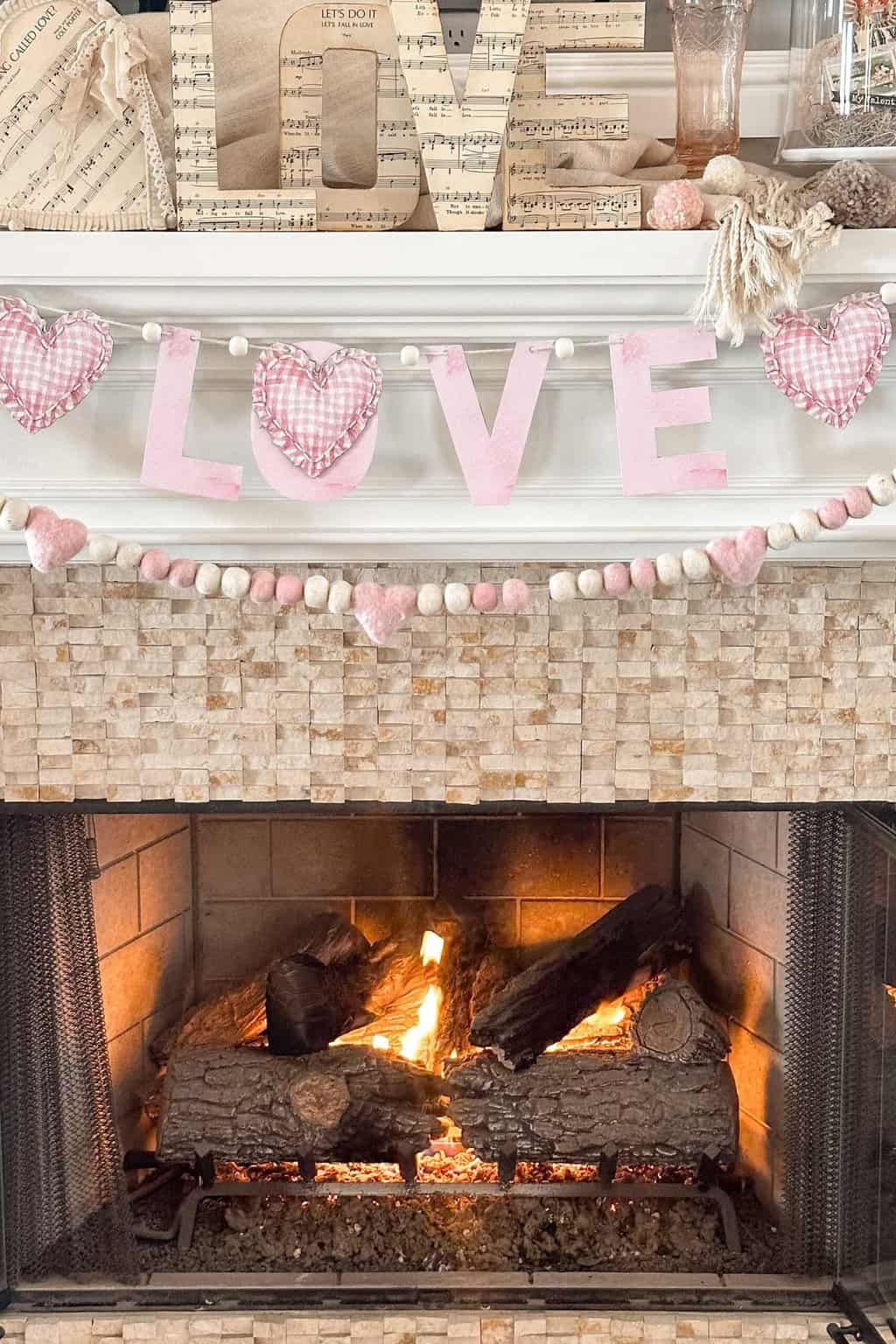 A cozy fireplace decorated with a "LOVE" theme features a garland of pink and white felt hearts, with "LOVE" spelled out in fabric letters. The mantel holds music sheet-covered letters spelling "LOVE" and other decorative items, complemented by a free printable Happy Valentine's Day banner.