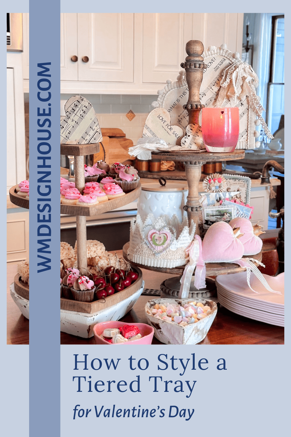 A valentines tiered tray decor features heart-shaped items, pink and red sweets, a candle, decorative books, and dishes, all arranged in a cozy kitchen with white cabinets as the backdrop.