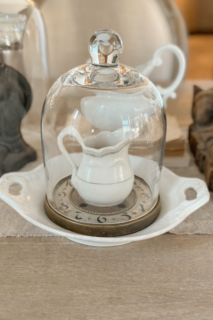 A small white ironstone pitcher under a cloche sitting on an old clock face.