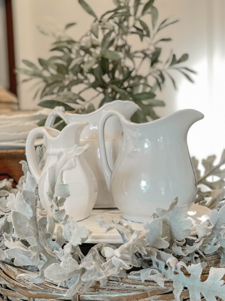 Three white ironstone pitchers are sitting in a wood basket with faux greenery for a table display for winter.