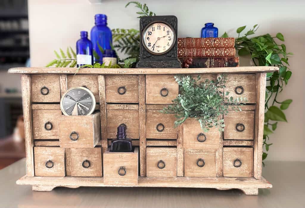 Winter Decorations (Not Christmas)-A rustic wooden box with drawers sits on a desk decorated for winter with faux greenery, vintage clocks, old books, and bright blue glassware.