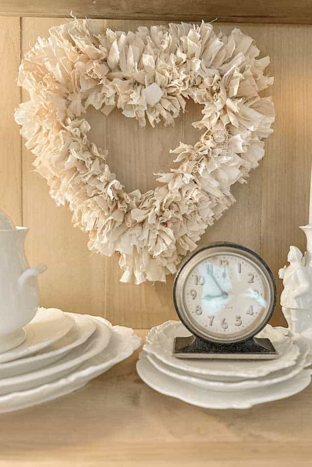 A decorative white heart-shaped wreath made of fabric hangs on a wooden wall. Below it, a vintage clock, some white plates, a porcelain tureen, and a white candle holder are arranged on a wooden shelf. This charming setup provides rustic and cozy Valentine's Day dinner table decoration ideas.