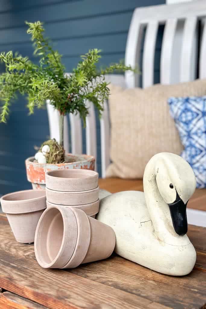 Small details of a wood goose, a topiary tree, and beautiful pots are displayed on the top of a coffee table on the front porch for budget-friendly decor.