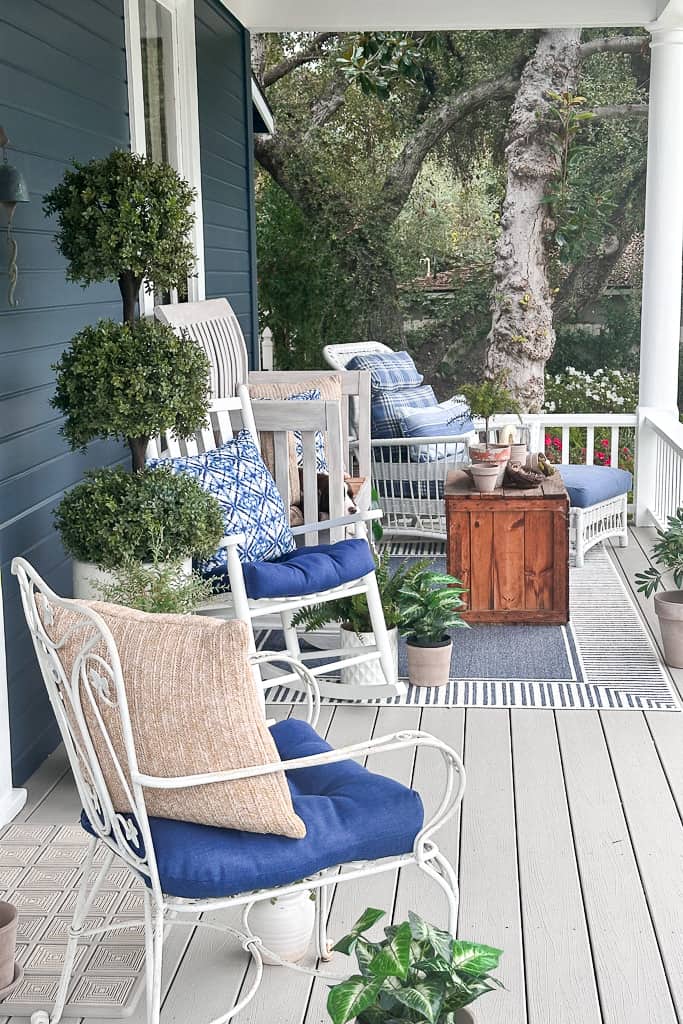 A cozy front porch with blue walls features white rocking chairs with blue cushions, a wooden coffee table, potted plants, and a tree in the background. The porch is decorated with a mix of natural and nautical elements, creating a relaxing outdoor space.