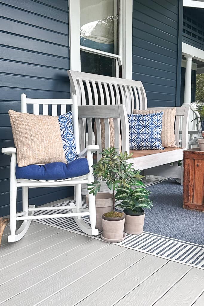 Small faux green plants are sitting next to a white rocking chair on the front porch. 