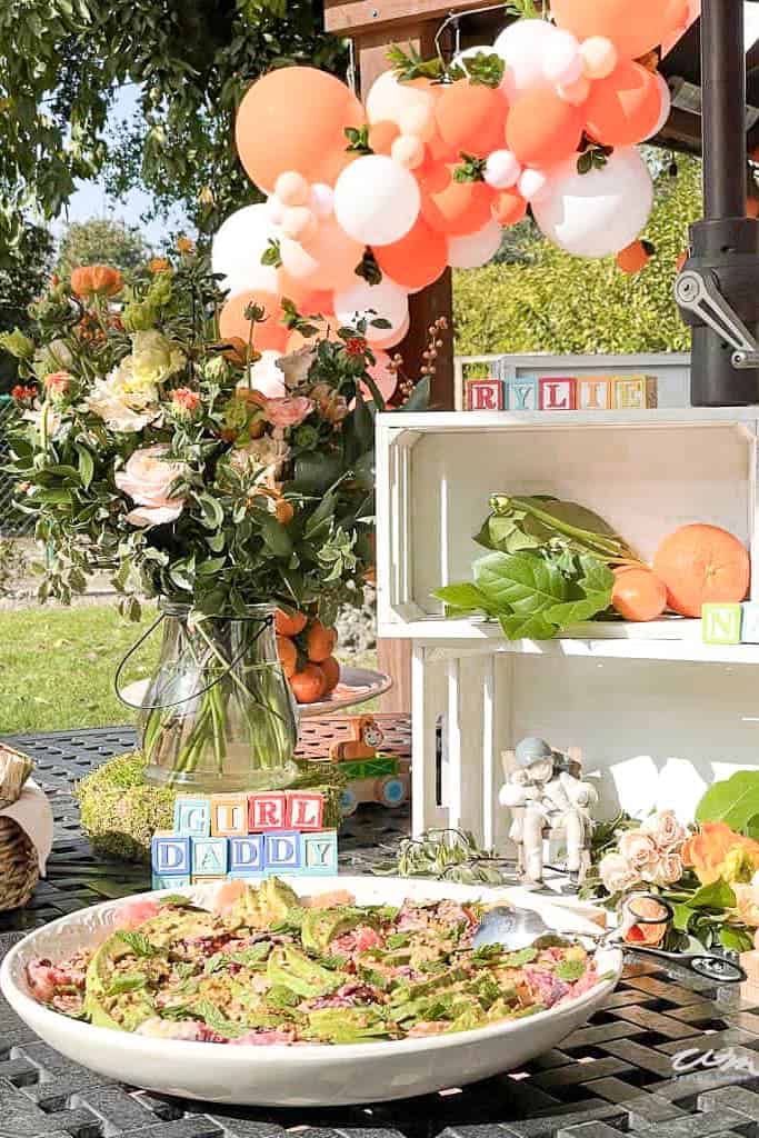 A plate of avocado salad sits on the table with baby blocks, fresh flowers, and a balloon arch.