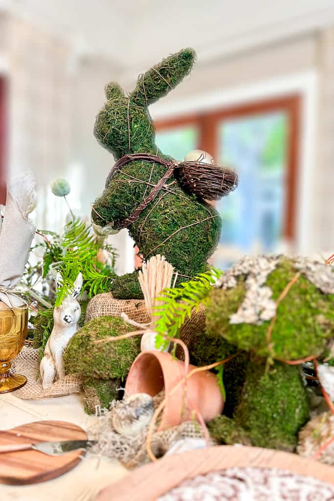 A centerpiece features a moss-covered rabbit figurine with a small basket on its back, surrounded by greenery, mushrooms, a small ceramic bunny, and pink flower pots, all on a beige tablecloth. The background shows a bright room with large windows.