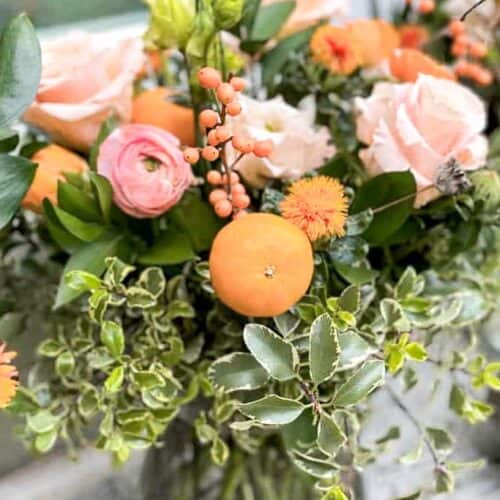 A vibrant floral arrangement in a glass vase featuring a mix of orange and pink flowers, including roses and berries, surrounded by lush green leaves and foliage. The stunning floral arrangement is set on a light-colored surface with a blurred outdoor background.