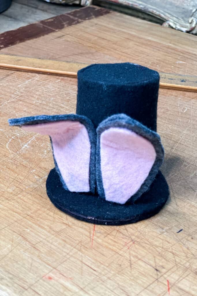 Attaching the top of the top hat over the ends of the ears. Final black felt top hat with two grey and pink ears.