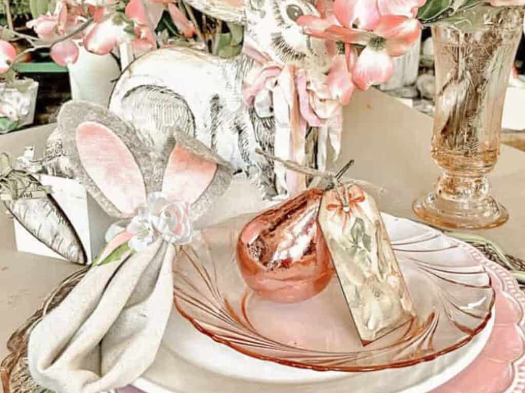 An Easter place setting with a pink glass plate, a pink metallic pear with a personalized name card attached. Eater rabit DIY Ears made out of felt.