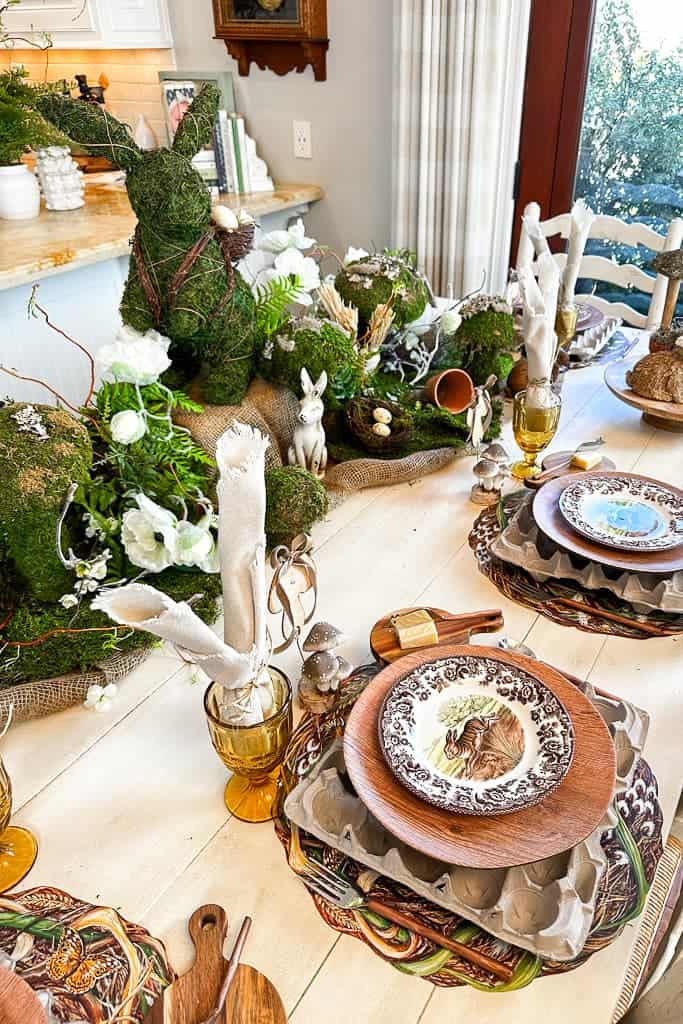 A table setting with an elaborate Easter theme. The table is adorned with moss-covered bunny decorations, white flowers, and rustic elements. Each place setting features a patterned plate, wooden charger, rolled cloth napkin, and an amber-colored glass.